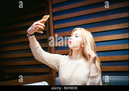 Junge blonde Frau in einem gestrickten Pullover, eine selfie Foto auf einem Smartphone, an einem Tisch in einem Cafe sitzen, Zeit allein, Internet Kommunikation Stockfoto