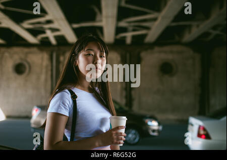 Süße junge Asiatin mit einem Glas zum mitnehmen, geht in einem städtischen Umfeld Stockfoto