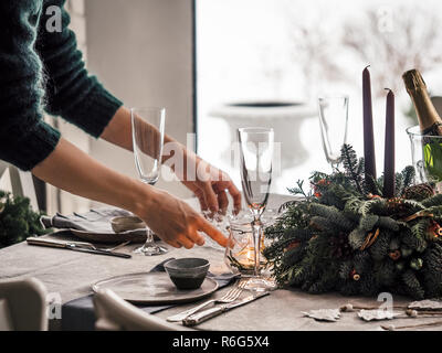 Weibliche Hände legen Sie die Tabelle für das Weihnachtsessen im Wohnzimmer. Schneereiche Winter im Fenster im Hintergrund. Schöne serviert Tabelle mit Dekorationen, Kerzen, Tannenzweigen. Urlaub Stockfoto