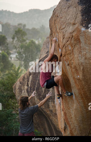 Ein männlicher Kletterer Praktiken, seine Fähigkeiten, Grip und Stärke Klettern ein Boulder mit einem Freund unter ihm Spotting für Sicherheit. Stockfoto