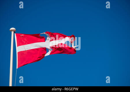 Ein zerschlagener zerrissen und zerrissene Flagge von Dänemark (Rot mit weißem skandinavischen Kreuz, die sich an den Rändern des Flag erweitert; Der vertikale Teil des Kreuzes ist die Hoist Seite verschoben), Fliegen auf einem Fahnenmast mit einem tiefblauen Himmel hinter Stockfoto