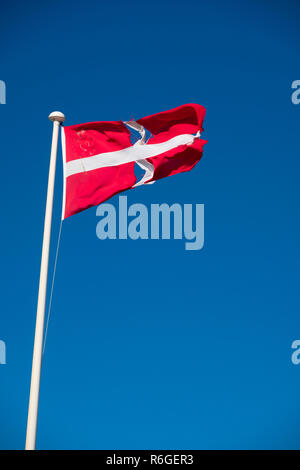 Ein zerschlagener zerrissen und zerrissene Flagge von Dänemark (Rot mit weißem skandinavischen Kreuz, die sich an den Rändern des Flag erweitert; Der vertikale Teil des Kreuzes ist die Hoist Seite verschoben), Fliegen auf einem Fahnenmast mit einem tiefblauen Himmel hinter Stockfoto