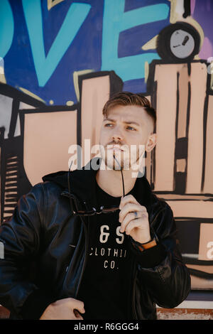 Junger Erwachsener Mann mit Sonnenbrille außerhalb in der kühlen Jahreszeit Downtown Urban Street Art District Sitzen vor dem pulsierenden bunten Graffiti Art Wall Stockfoto