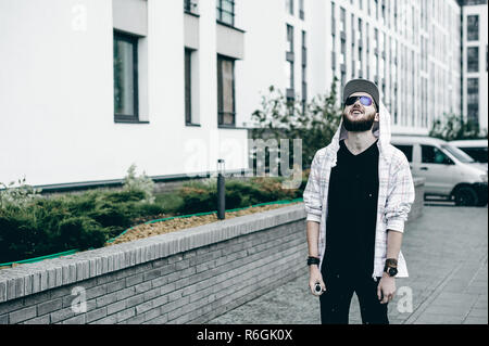 Junger Mann mit Bart in hell karierten jacke mit Kapuze in einem Hut und Sonnenbrille frei herumlaufen Stadt raucht eine e-Zigarette Stockfoto