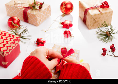 Prozess Verpackung Weihnachtsgeschenke. Weiße Holztisch mit roten Verzierungen, Kugeln, Kerzen, Sterne, weibliche Hände Geschenk rot Goldband schmücken. Neues Jahr, winter Konzept. Stockfoto