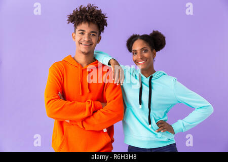 Foto von jugendlich afrikanische amerikanische Paar in bunten Kleidern lächelnd, während zusammen über violette Hintergrund stehen isoliert Stockfoto