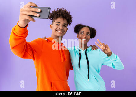 Foto von Happy african american Paar tragen bunte Sweatshirts unter selfie auf dem Smartphone über violett Hintergrund isoliert Stockfoto