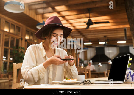 Portrait von zufriedenen Frau mit Hut fotografieren Essen auf Handy während der Sitzung in stilvollen Café Stockfoto