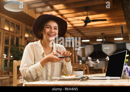 Portrait von Frau Hut tragend fotografieren Essen auf Handy während der Sitzung in stilvollen Café Stockfoto