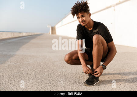 Starker junger afrikanischer Mann in Sportkleidung band seine Schnürsenkel am Strand gekleidet Stockfoto