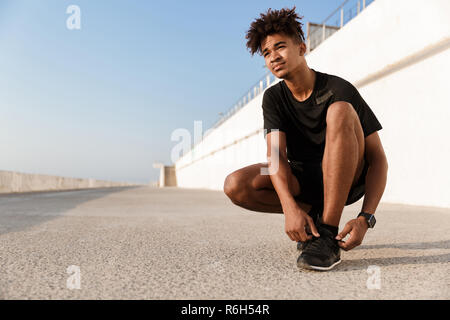 Selbstbewussten jungen afrikanischen Mann in Sportswear band seine Schnürsenkel am Strand gekleidet Stockfoto