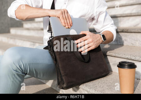 Foto von Geschäftsmann 30 s Sonnenbrille tragen, Laptop in der Tasche beim Sitzen auf der Treppe auf die Straße Stockfoto