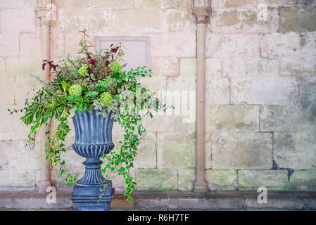 Stein vase Pflanzmaschine mit Fustian grün Blumen und Kaskadierung grün Efeu und Blätter. Grauen Pflastersteinen. Wunderschönes Design vor medevial Wand mit c Stockfoto