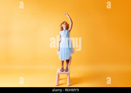 Ein kleines Kind Mädchen in einem blauen Kleid steht auf einem Stuhl und Maßnahmen ihrer Höhe vor dem Hintergrund der gelben wand. Konzept der Entwicklung, Stockfoto