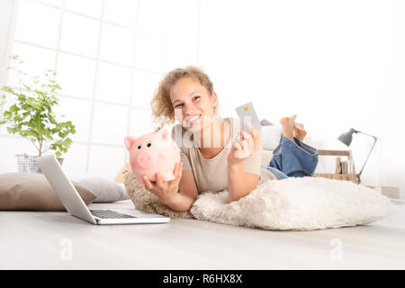 Junge lächelnde Frau mit Kreditkarte und Sparschwein, mit Laptop Computer online shopping sparen Konzept, liegen auf Wohnzimmer Holzboden in Co Stockfoto