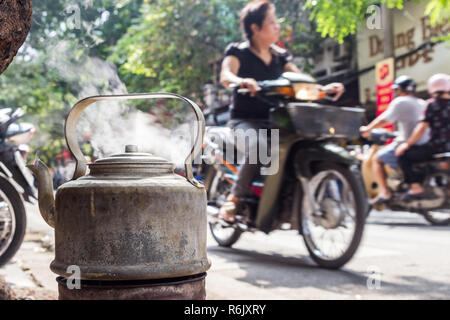 Kochendes Wasser in alten Wasserkocher aus Metall auf der Straße in Hanoi, Vietnam. Hintergrund verschwommen, nicht erkennbare Frau auf dem Motorroller. Stockfoto