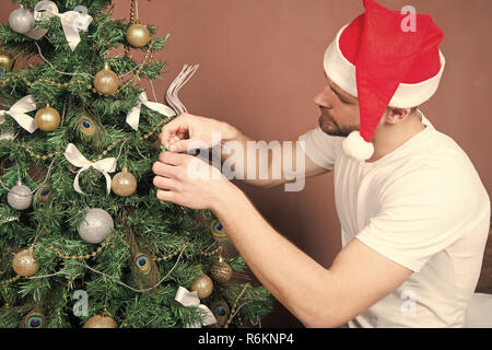 Mann in santa hut Weihnachtsbaum dekorieren mit Kugeln, Schleifen, Girlande. Weihnachten, Neujahr, Silvester, urlaub Vorbereitung, Feier. Festliche Dekorationen und Verzierungen Konzept Stockfoto