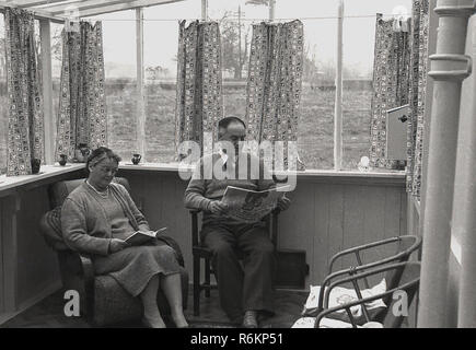 1970er Jahre, ein Rentnerehepaar sitzen in ihren kleinen Holz gerahmt Wintergarten lesen, der Mann, der eine Zeitung und die Dame ein Buch, England, UK. Stockfoto