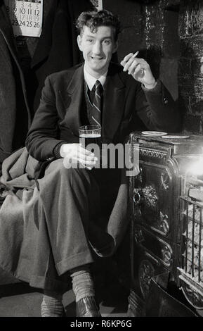 1940 s, historischen, ein junger Mann, der in seinen Zwanzigern, trägt eine Jacke und Krawatte, in einer Kneipe sitzen, die von einem Brand, mit einem Pint Bier und einer Zigarette. Stockfoto