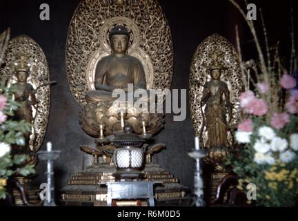 Farbfoto, von einem niedrigen Winkel, in dem ein fein geschnitzten und vergoldeten Buddha, sitzend auf einem Lotus Blume, mit einem flammenden Mandorla im Hintergrund, und seine Hände zusammen in einem meditativen Mudra, mit Twin attendant Figuren flankiert, die jeweils in einer Mandorla, und mit Blumen, Kerzen und/oder eine Pfanne im Vordergrund sichtbar, wahrscheinlich in einem Japanischen, buddhistische Tempel in der Mitte des Zwanzigsten Jahrhunderts, 1965 fotografiert. () Stockfoto