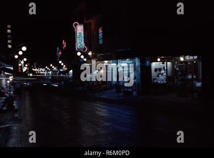 Farbe, vintage Foto von einem Market Street, in der Nacht fotografiert, mit Handtaschen und Kleidung sichtbar durch beleuchtete Schaufenster Links, Leuchtreklame mit Japanischen schreiben, und die Reihen der Triple-bulbed Lampe Beiträge Kennzeichnung Kurve der Straße, in Japan, 1965. () Stockfoto