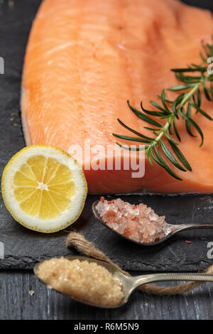 Roher Lachs filet auf schwarzem Schiefer Platte und Zutaten für die Herstellung von Graved Lachs. Salz, Zucker, Zitrone und Rosmarin im Hintergrund für trockene Heilung Marinade, da Stockfoto