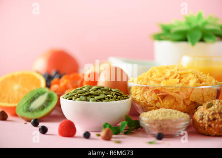 Cornflakes mit Milch, Kürbiskerne, frische Beeren, Joghurt, gekochte Eier, Nüsse, Obst, Orange, Banane, Pfirsich für Frühstück auf rosa Hintergrund. Kopieren Sie sp Stockfoto