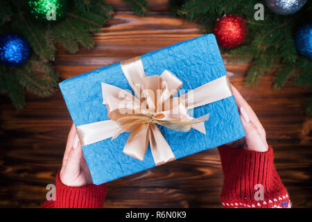 Ansicht von oben. Woman's Hände mit Geschenk in blaues Papier mit Goldband gewickelt. Holz- vintage Tabelle. Schöne Ferien Hintergrund mit grünen bran Fichte Stockfoto