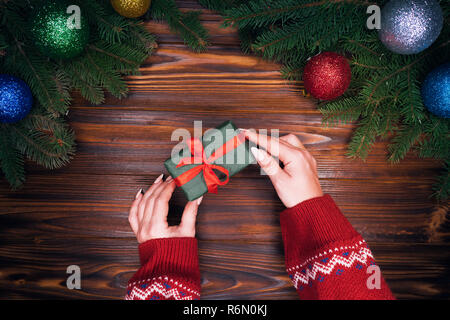 Weibliche Hände legt Geschenk in Emerald Papier mit rotem Band auf retro Holztisch mit Weihnachtsschmuck - grüne Zweige Fichte gewickelt, Weihnachten ba Stockfoto