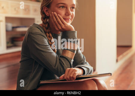 Kleines Mädchen sitzt auf dem Boden mit ihrem Buch und Wegsehen denken. Mädchen mit Geschichte Buch weg schauen und sich vorstellen. Stockfoto