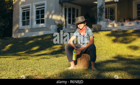 Wenig cowboy sitzen draußen im Hinterhof mit Wasserpistole. Junge Junge, tragen, Cowboy Hut mit Wasser squirt Gun. Stockfoto