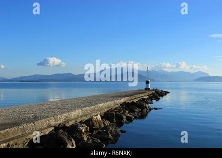 Leuchtturm am Ende der Pier am Koronisia Dorf in Ambracian Golf, Epirus Griechenland, ruhigen, blauen Meer, Wolken im Himmel und Blick auf die Berge und Hügel Stockfoto