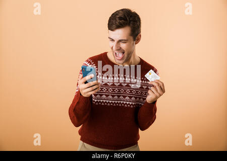 Portrait ein fröhlicher junger Mann in Pullover stehend über beigen Hintergrund isoliert gekleidet, Mobiltelefon, Kreditkarte Stockfoto