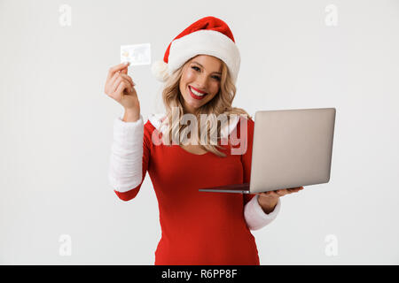 Portrait von freudiger blonde Frau in Rot neues Jahr Kostüm auf weißem Hintergrund gekleidet, mit Laptop, mit Kreditkarte Stockfoto