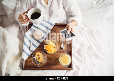 Ein Mittelteil der Frau im Bett lag, mit Kaffee und Frühstück drinnen in der Morgen in einem Schlafzimmer, mit Smartphone. Eine Ansicht von oben. Stockfoto