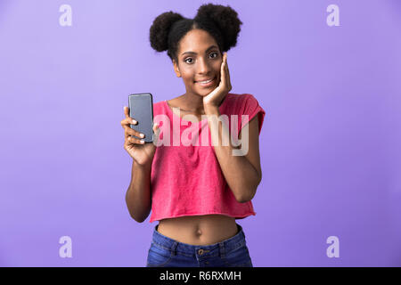 Foto von überrascht afrikanische amerikanische Frau und lächelnd Mobiltelefon über violette Hintergrund isoliert Stockfoto