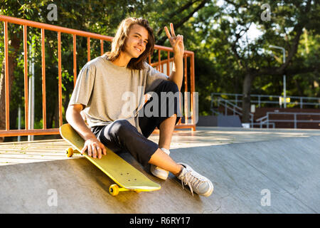 Foto männlicher Jugendlicher 16-18 in Freizeitkleidung auf Rampe mit Skateboard, Skate Park während der sonnigen Sommertag Stockfoto