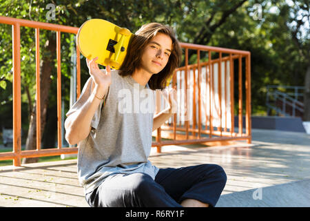 Foto von jugendlich Kerl 16-18 in Freizeitkleidung auf Rampe in Skate Park und halten Skateboard über die Schultern während der sonnigen Sommertag Stockfoto