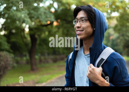 Bild von positiven asiatischer Mann in Freizeitkleidung und Brillen lächelnd bei einem Spaziergang durch den Green Park