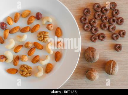 Mandeln, Haselnüsse, Walnüsse, Cashewnüsse auf einem weissen Teller, drei ganze Walnüsse, knackige Vollkornprodukte, Hafer auf hellem Holztisch. Gesund Stockfoto