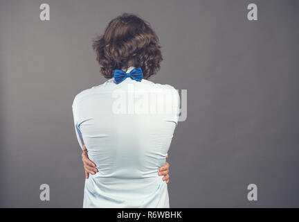 Junger Mann im blauen Hemd und Fliege schmiegt sich auf grauem Hintergrund. Ansicht von hinten. Seine Hände liegen auf der Taille. Er muss angenommen zu werden. Egoistische gefühl, dich zu lieben. Positive Emotion. Platz kopieren Stockfoto