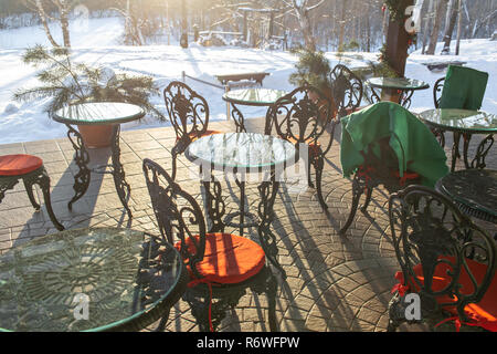 Gusseiserne Tische und Stühle mit Kissen und eine Decke auf Restaurant Terrasse. Winterlandschaft von frostigen Bäumen, weißer Schnee und blauer Himmel. Ruhigen winter natur im Sonnenlicht in Park