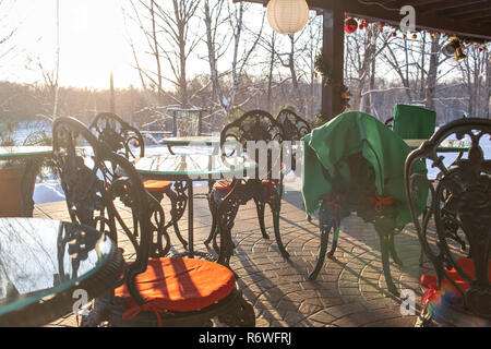 Gusseiserne Tische und Stühle mit Kissen und eine Decke auf Restaurant Terrasse. Winterlandschaft von frostigen Bäumen, weißer Schnee und blauer Himmel. Ruhigen winter natur im Sonnenlicht in Park