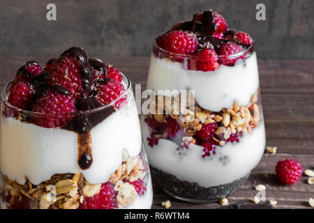 Himbeer Joghurt Parfait in Gläsern mit Schokolade, Müsli und Chia Samen für gesundes Frühstück auf rustikalen Holzmöbeln Hintergrund. Nach oben Schließen Stockfoto