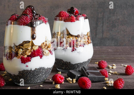 Himbeer Joghurt Parfait in Gläsern mit Schokolade, Müsli und Chia Samen für gesundes Frühstück auf rustikalen Holzmöbeln Hintergrund Stockfoto