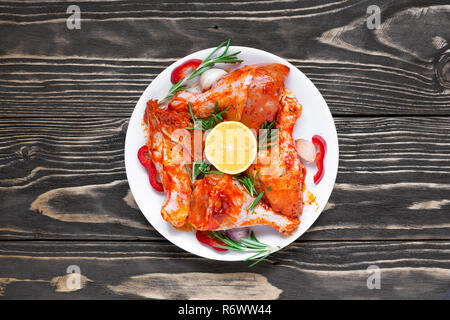Roh marinierte Hähnchenschenkel mit roten pikanter Sauce und Kräuter in eine Platte auf Holz- Hintergrund. Vorbereitung zum Kochen und Grillen, Ansicht von oben Stockfoto
