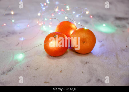 Bild von Mandarinen auf Schnee mit Girlande. Mandarinen und Girlanden. Weihnachtliche Stimmung. Selektive konzentrieren. Weihnachten oder Neujahr. Frische reife Mandarinen. Weihnachten noch Leben Stockfoto