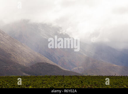 Dramatische Berglandschaft mit Licht durch Regenwolken auf nasse Oberfläche, unten ist ein Tal der Weinberge oder Weinreben Hex River Tal Stockfoto