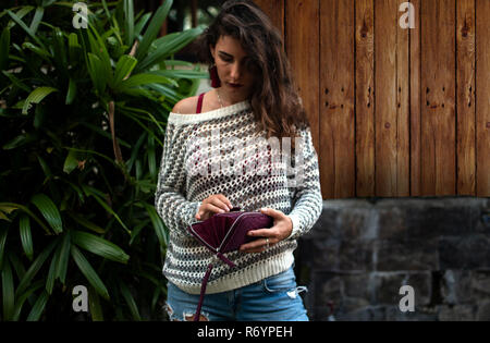 Frau hält einen lila python Geldbeutel in die Hände. Geld und Shoping. Die Frau Hände mit Tasche, Natur Hintergrund, Palmen Blätter, Holz, Jeans, Mode Stil Stockfoto