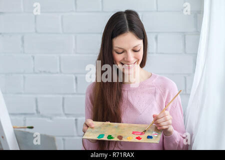 Schöne junge Frau malen ein Bild auf einer Staffelei. In der Hand einen Pinsel und Palette von Farben. Stockfoto